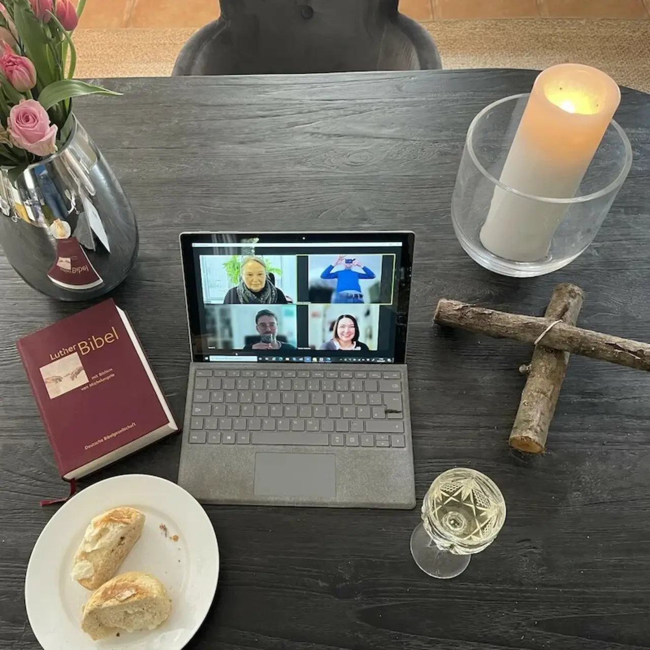 Tisch mit Blumen, einer Bibel, Brot, Wein, einem Kreuz aus Holz, einer Kerze und einem aufgeklappten Computer auf dem man mehrere Gesichter sieht.