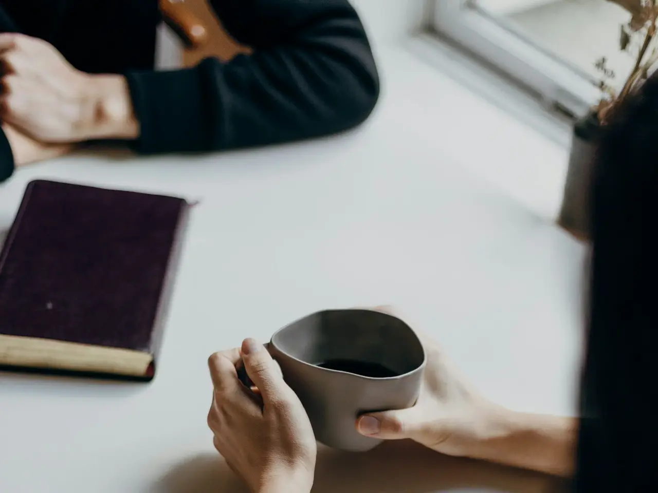 Unterarme von zwei Personen die an einem Tisch sitzen, eine Person hält eine Tasse in der Hand und auf dem Tisch liegt ein Buch.